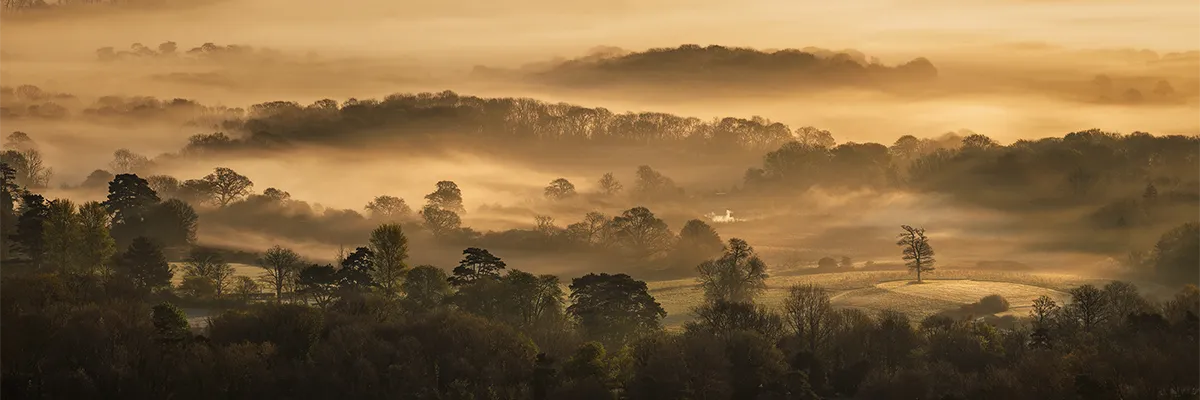 Golden morning mist drifting across layered countryside below Ditchling Beacon in East Sussex