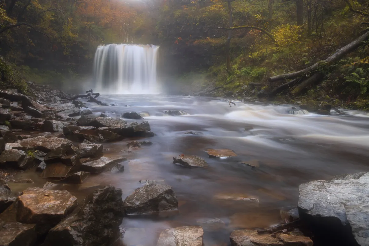 A wide waterfall cascading into a misty river surrounded by woodland in the Brecon Beacons, Wales